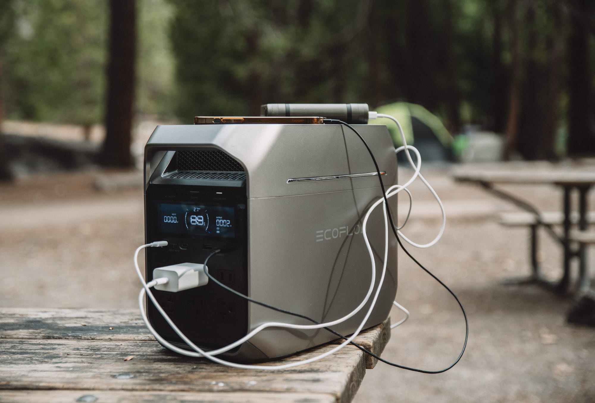 DELTA 3 Plus on the corner of a picnic table, white and black cables looping from its front ports and a white USB-C charger in an AC outlet, a phone and a small device on its top, tents and trees behind