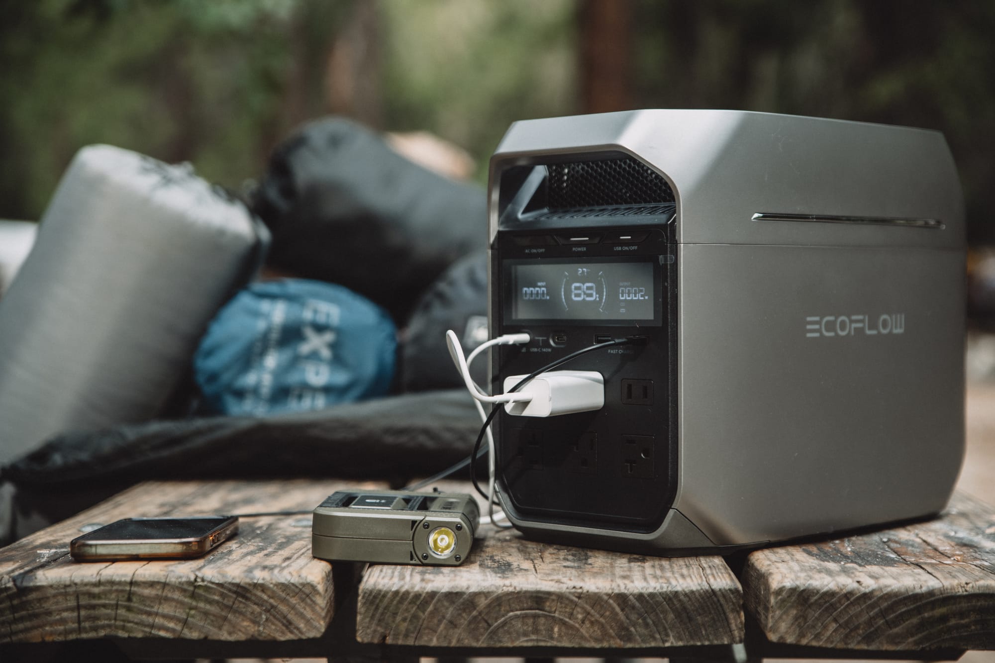 EcoFlow DELTA 3 Plus on a weathered campground picnic table, display lit, a white USB-C wall charger in one AC outlet and a cable in a USB port, a phone, a sleeping pad and stuff sacks beside it