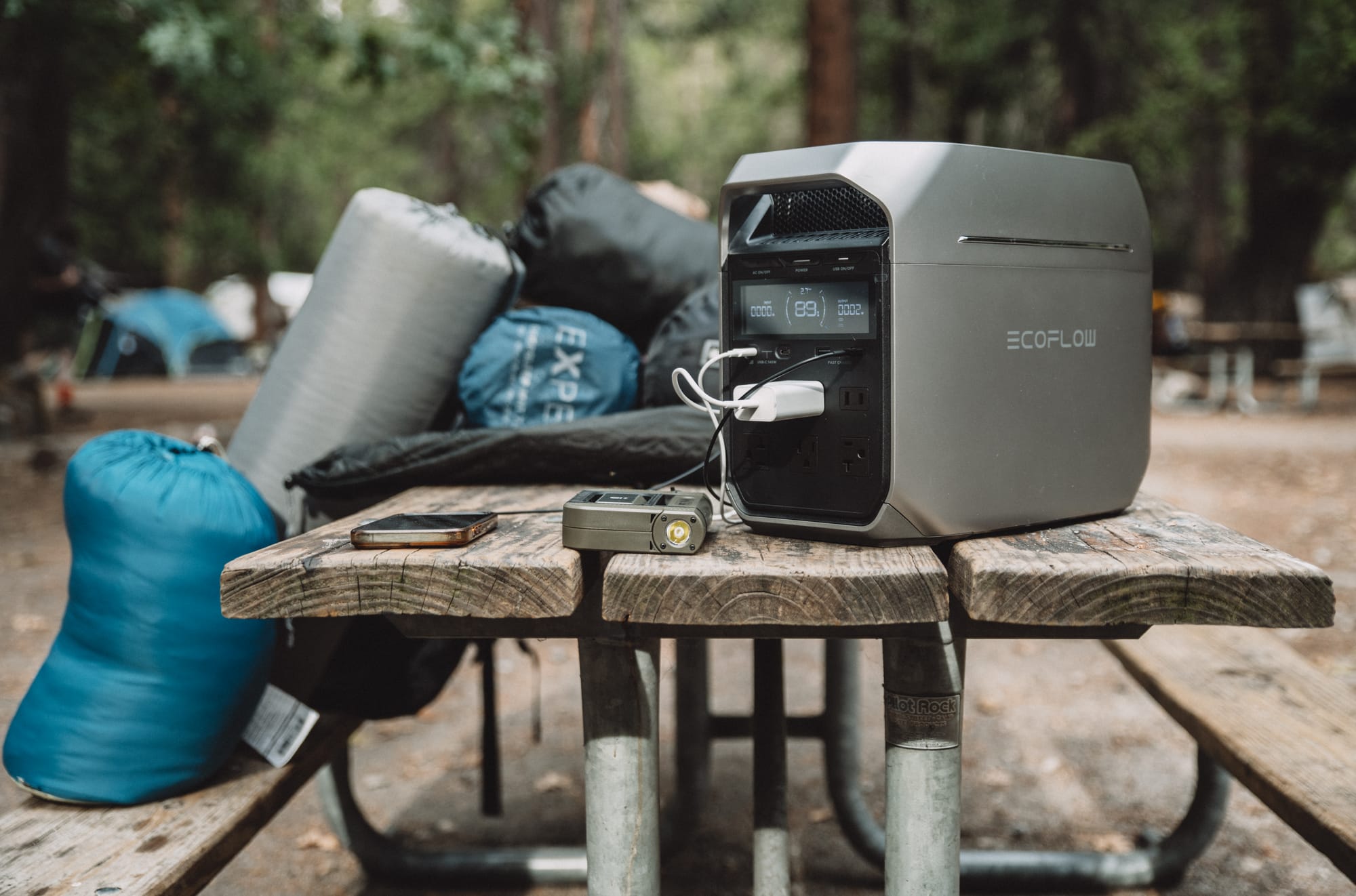 Wide view of a campsite: the DELTA 3 Plus at one end of a picnic table beside a rolled sleeping pad, stuff sacks and a blue sleeping bag, tents and pines behind