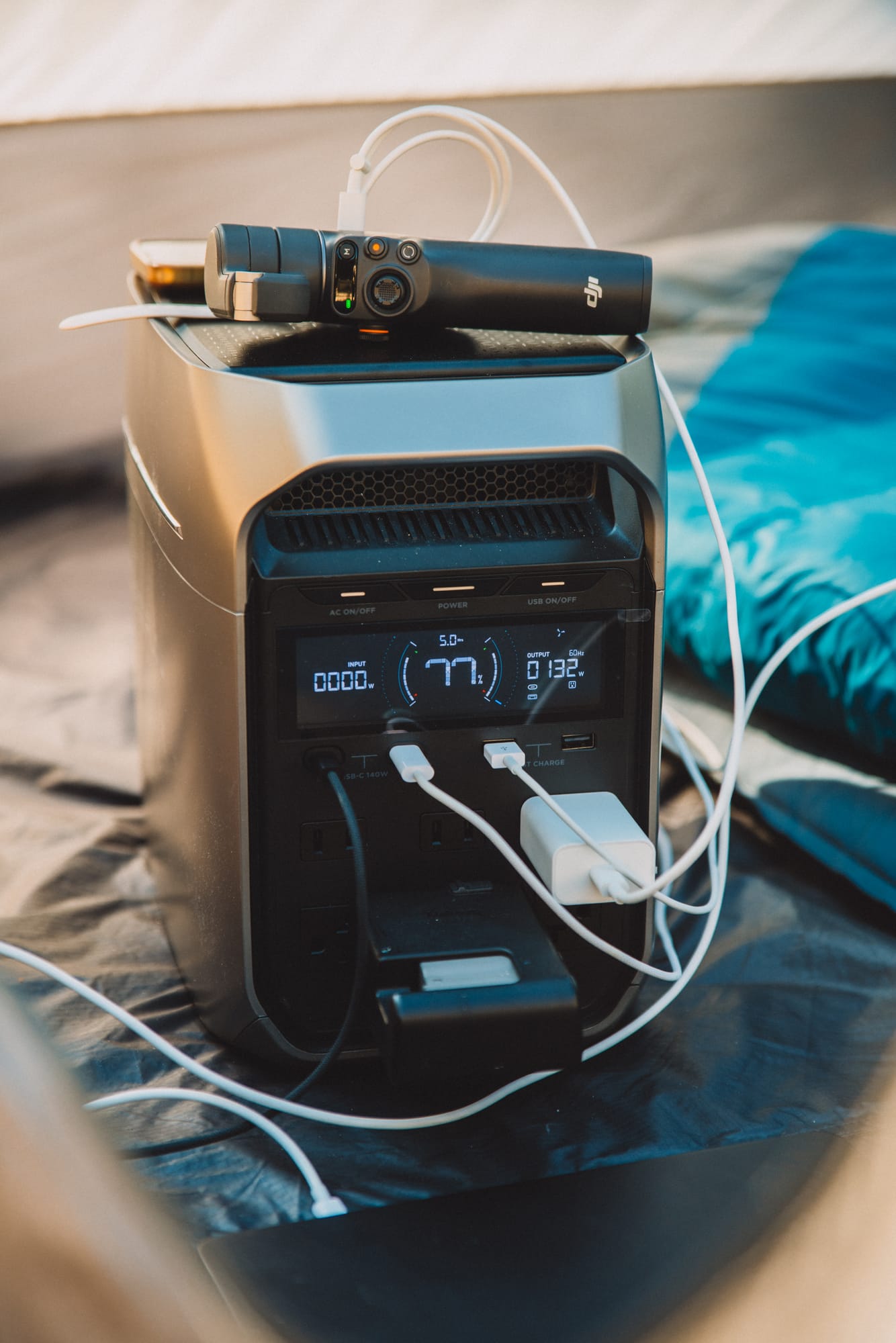 Close-up of the DELTA 3 Plus display reading input 0 W, 77%, 5.0 hours and output 132 W at 60 Hz, with cables in both USB-C ports and a USB-A fast-charge port and a wall charger in an AC outlet