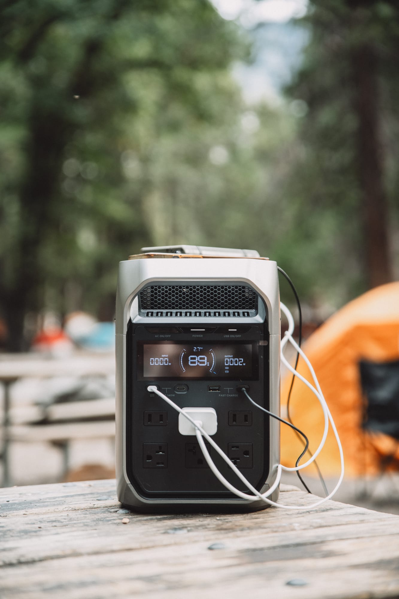 Front of the DELTA 3 Plus on a picnic table, display reading 89% with 2 W going out, a white USB-C wall charger in an AC outlet and cables in the USB ports, an orange tent behind