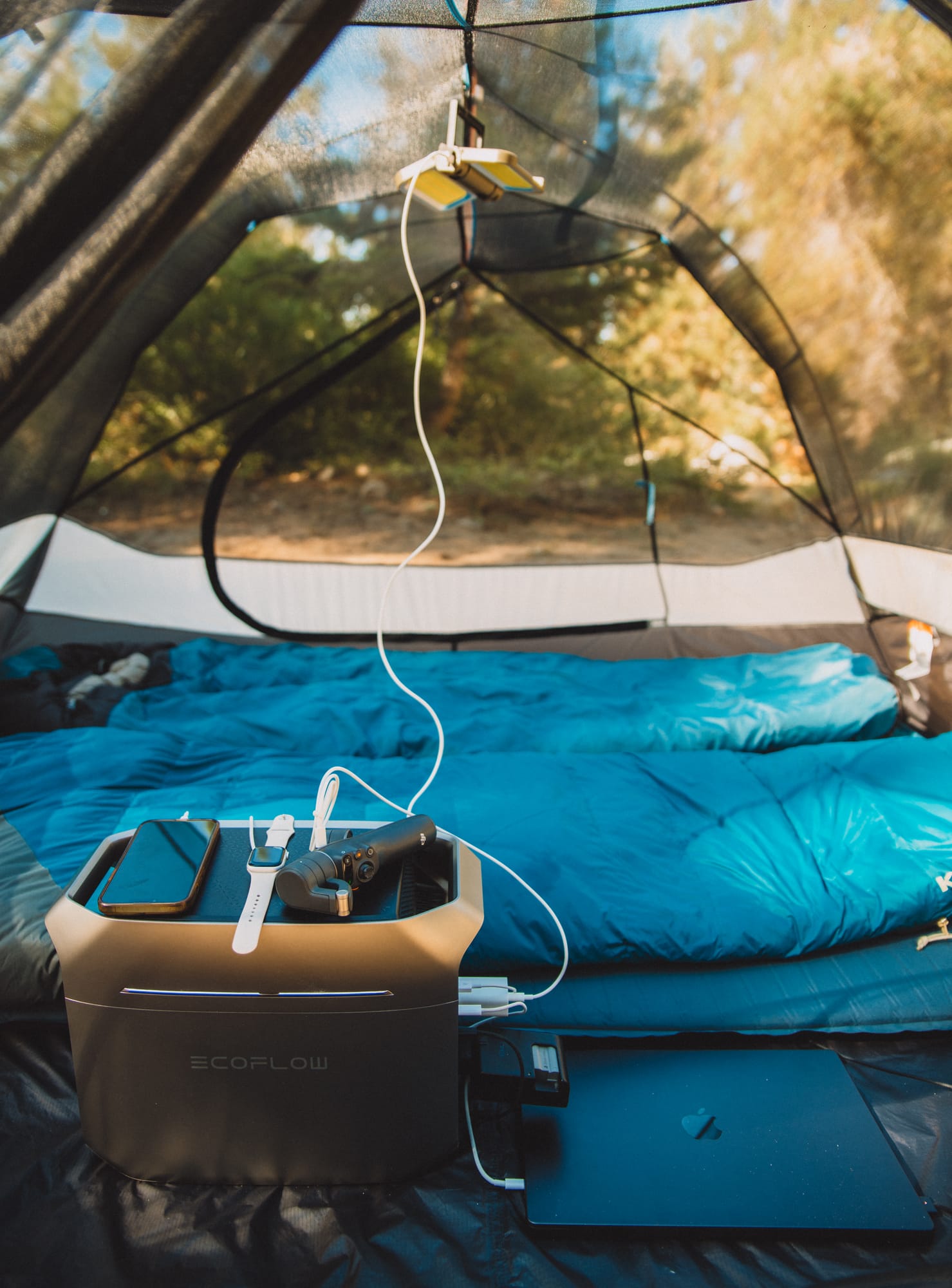 DELTA 3 Plus on a tent floor at the foot of two blue sleeping bags, a phone and a handheld camera on its top, a cable running up to a lantern clipped to the tent ceiling, a laptop lying beside it