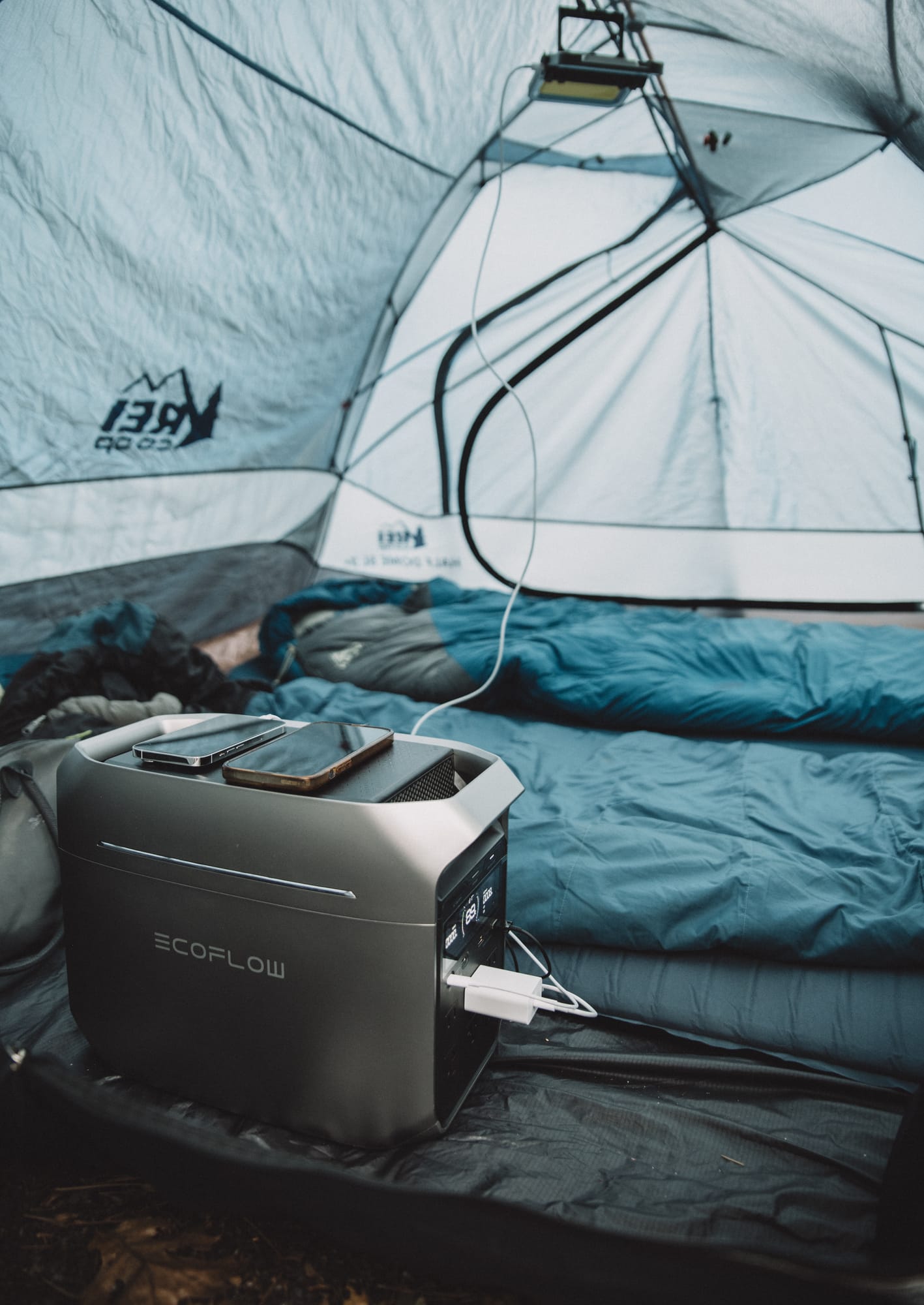 DELTA 3 Plus on a tent floor beside blue sleeping bags, two phones lying on its top, a USB wall charger in one outlet and a white cable running up to a lantern hung from the tent ceiling