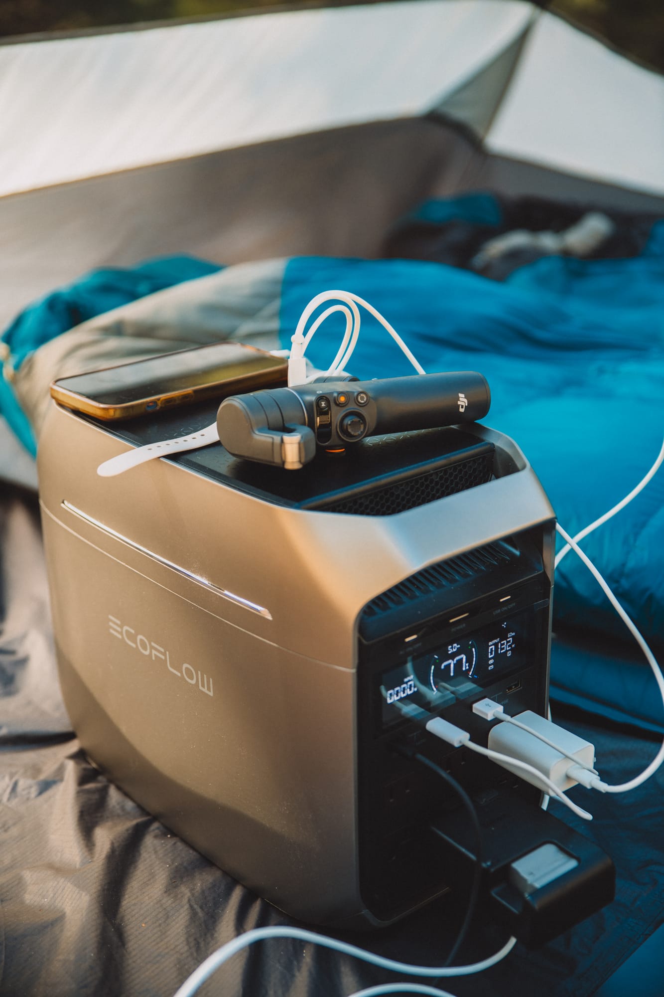 DELTA 3 Plus in a tent seen from above its front corner, a phone and a handheld gimbal camera lying on its top, cables running from the front USB ports and a wall charger in an AC outlet
