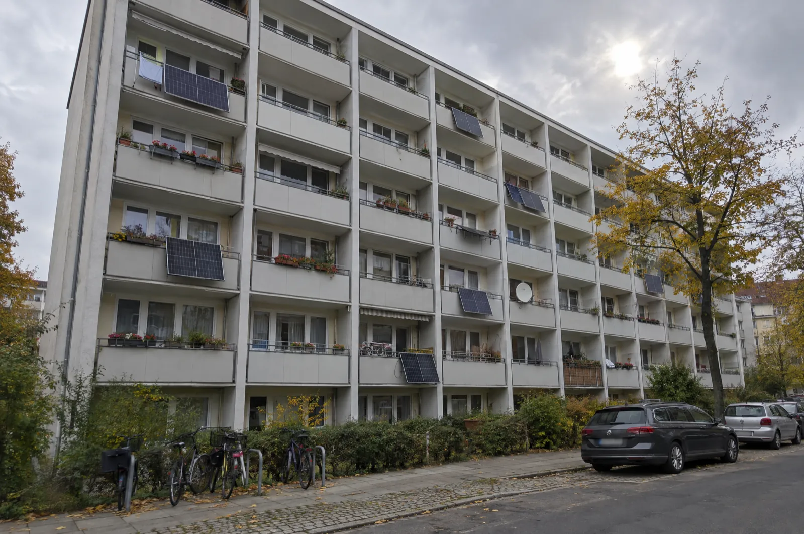 A six-story apartment block in Berlin on an overcast autumn day; several balconies have a single solar panel hung on the railing, among flower boxes and a satellite dish, with bicycles and parked cars on the street below.
