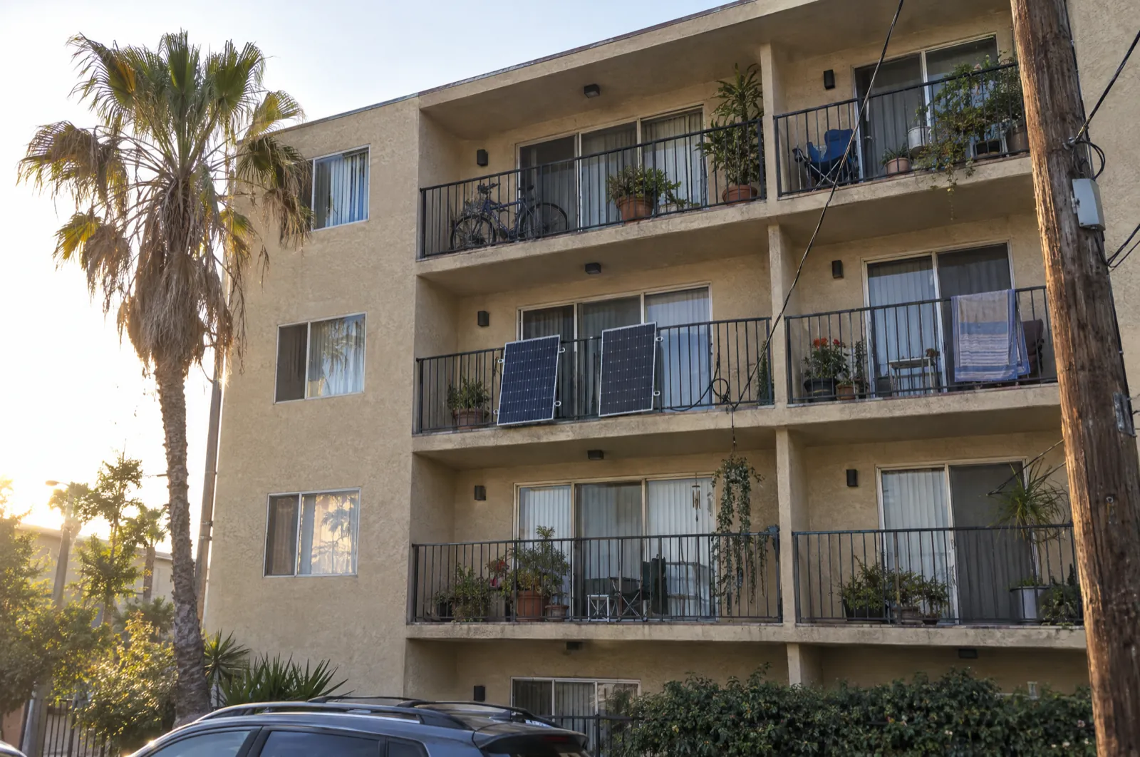 A plain four-story stucco apartment building with palm trees; on one third-floor balcony, two solar panels hang on the outside of the metal railing, a cable drooping between them.