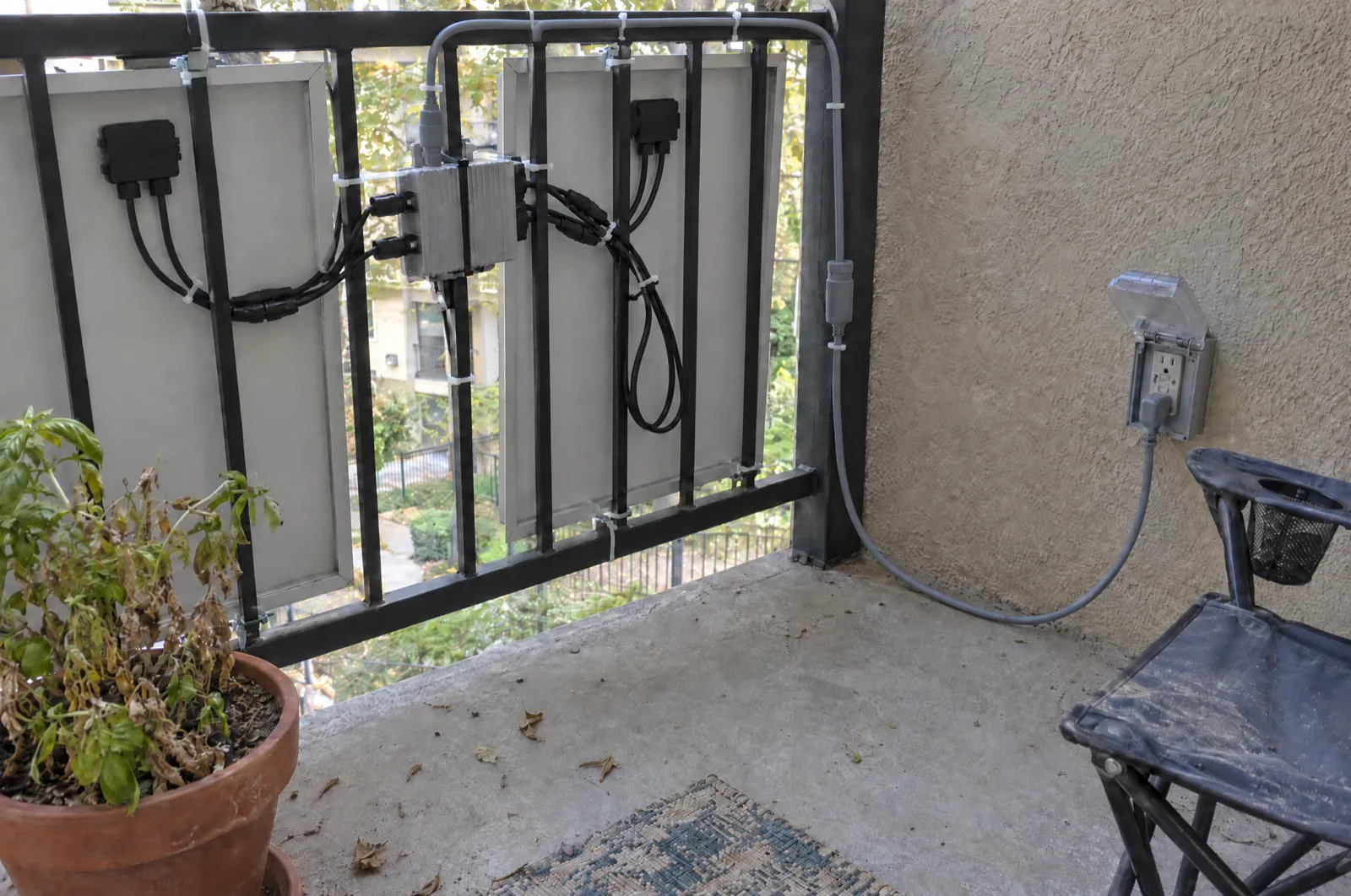 Seen from inside a balcony: the backs of two railing-mounted solar panels, a small grey microinverter zip-tied to the railing, and one grey cable running from the microinverter along the top rail, down the post and across the floor to a weatherproof outdoor outlet on the stucco wall.