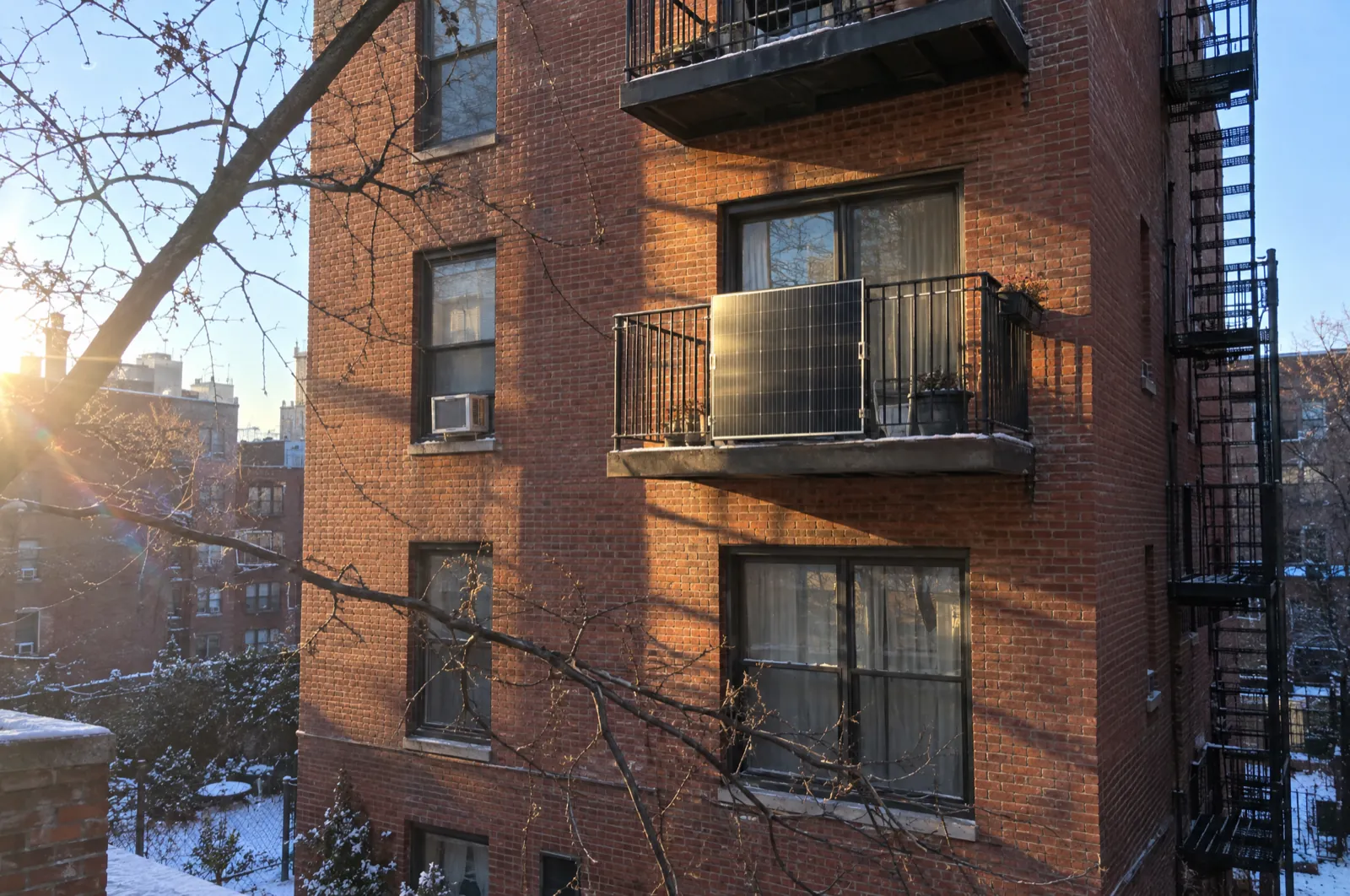 Late-afternoon December sun lights a single solar panel mounted on the iron railing of a brick apartment building, with old snow in the courtyard below and a bare tree branch in the foreground.
