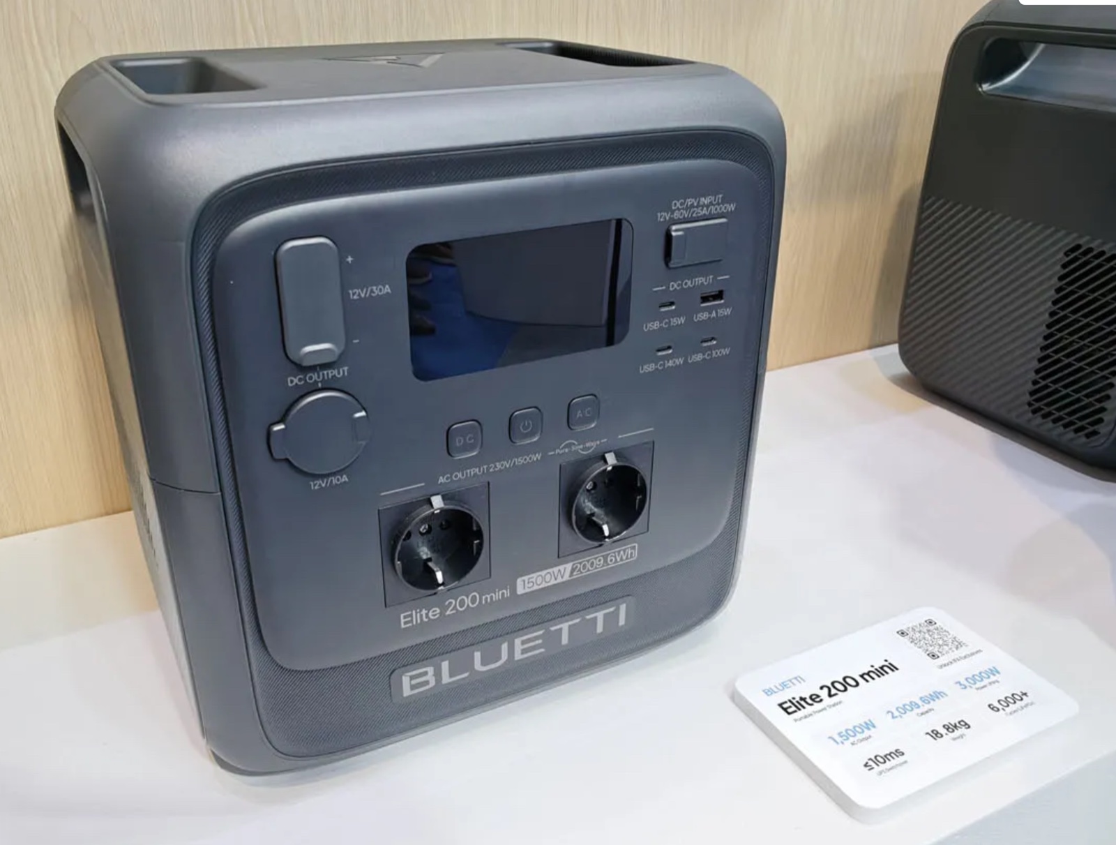 BLUETTI Elite 200 mini: a dark grey cube with a moulded handle channel, a recessed control panel carrying a 12V/30A terminal, a 12V/10A socket, a display, a four-port USB cluster and two Schuko outlets, with a badge reading 1500W 2009.6Wh, photographed beside its stand placard