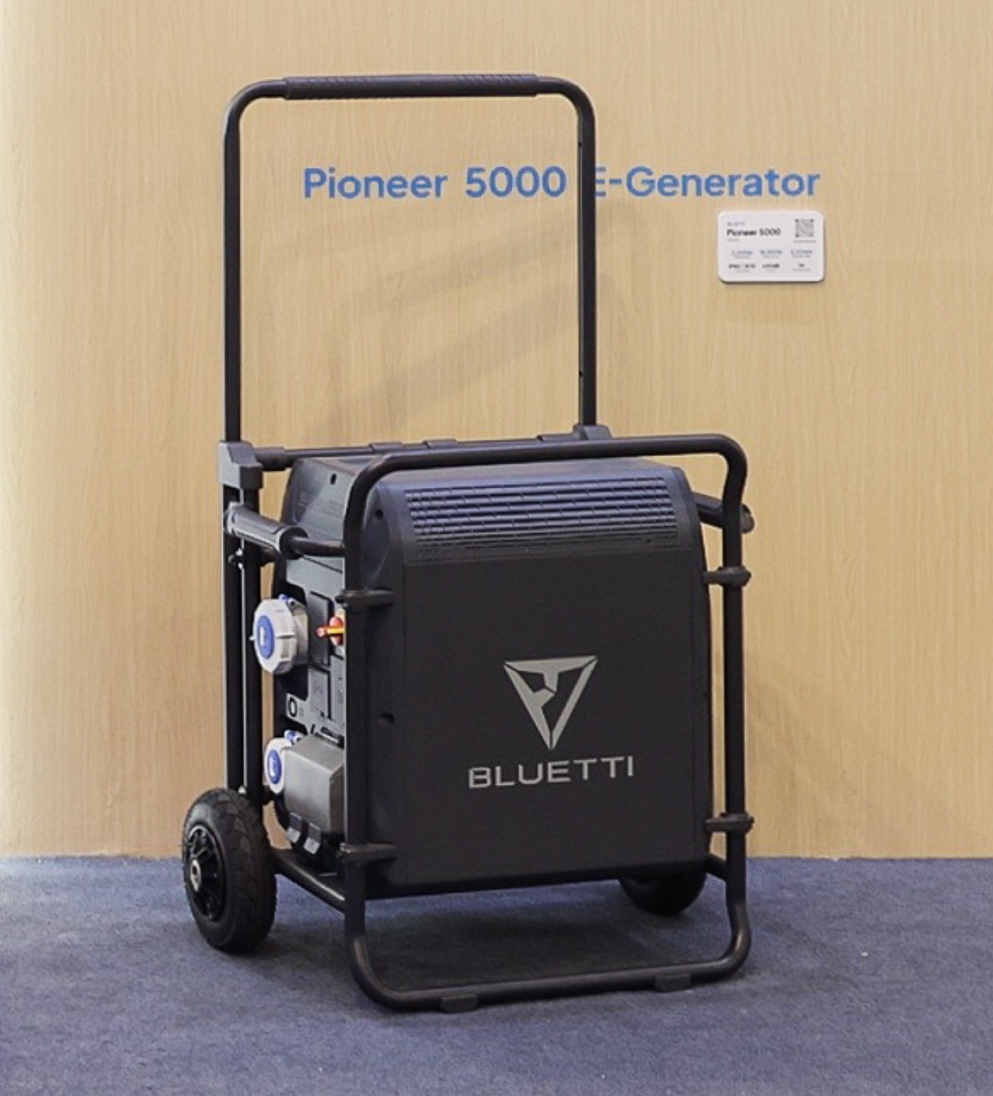 BLUETTI Pioneer 5000 E-Generator display unit at IFA Berlin 2026: a black cabinet inside a tubular roll-cage with a tall U-handle, two large knobby wheels and two blue industrial sockets on its left panel