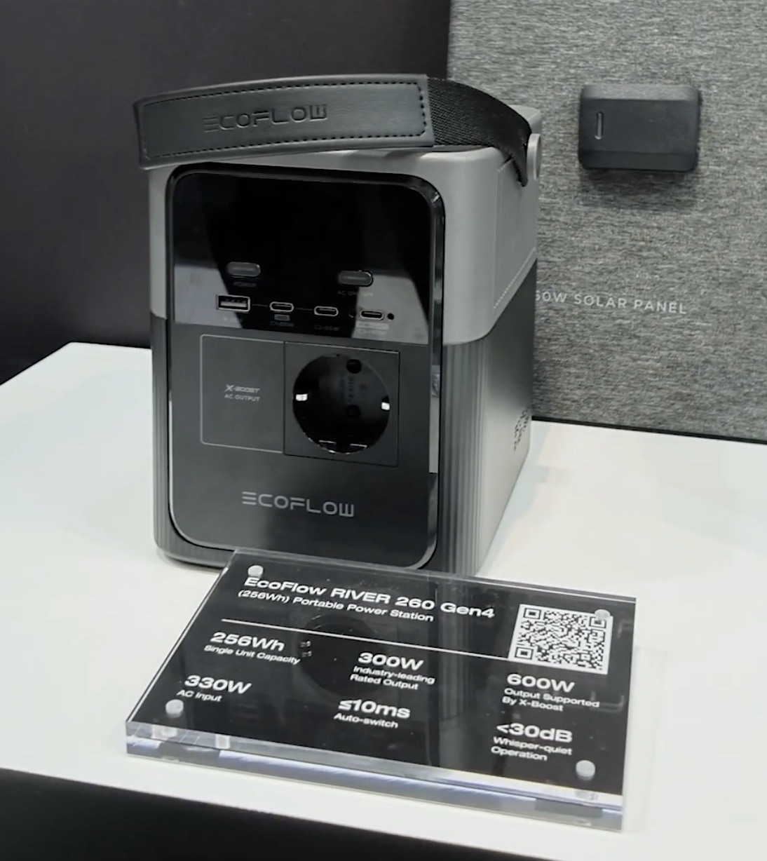 EcoFlow RIVER 260 Gen4: a compact grey power station with a webbing carry strap, a recessed black control panel carrying two buttons and a four-port USB row, and a single Schuko outlet in a bordered zone marked X-BOOST AC OUTPUT, photographed beside a placard listing its figures
